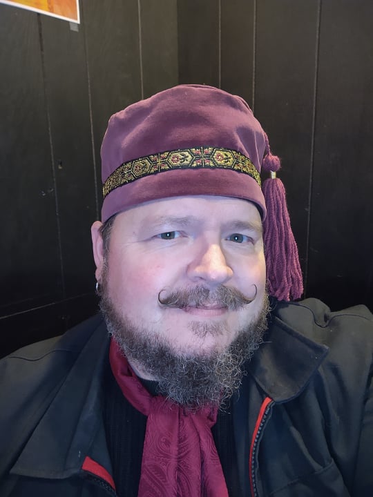 a round-faced man with a curly moustache wears a maroon velvet hat with a tassel on it